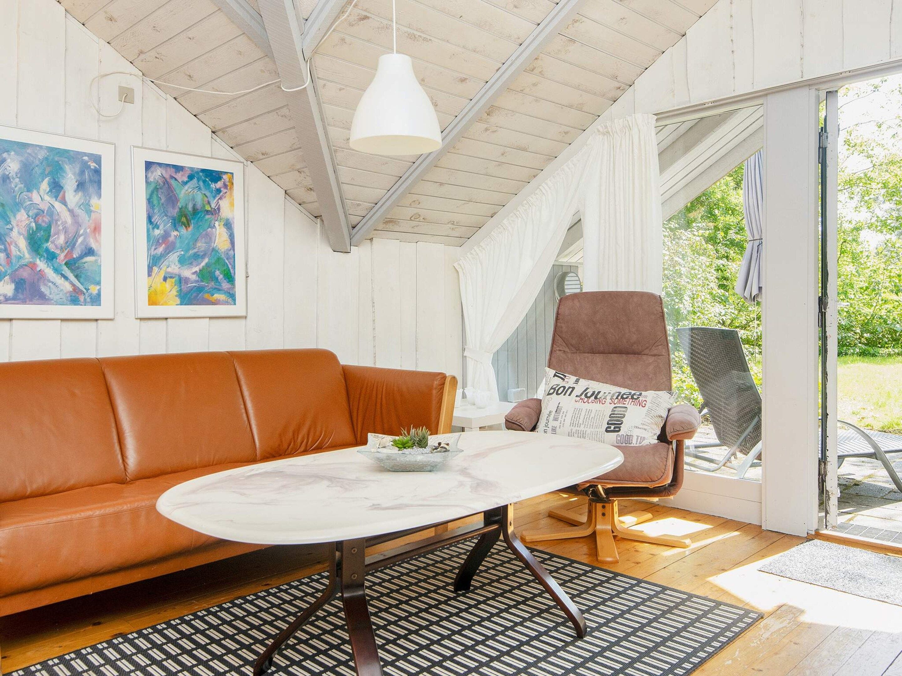 Una habitación con un sofá de cuero marrón, una mesa redonda con tapa blanca y una silla con cojín de periódico. Hay dos cuadros abstractos en la pared y un ventilador de techo blanco.