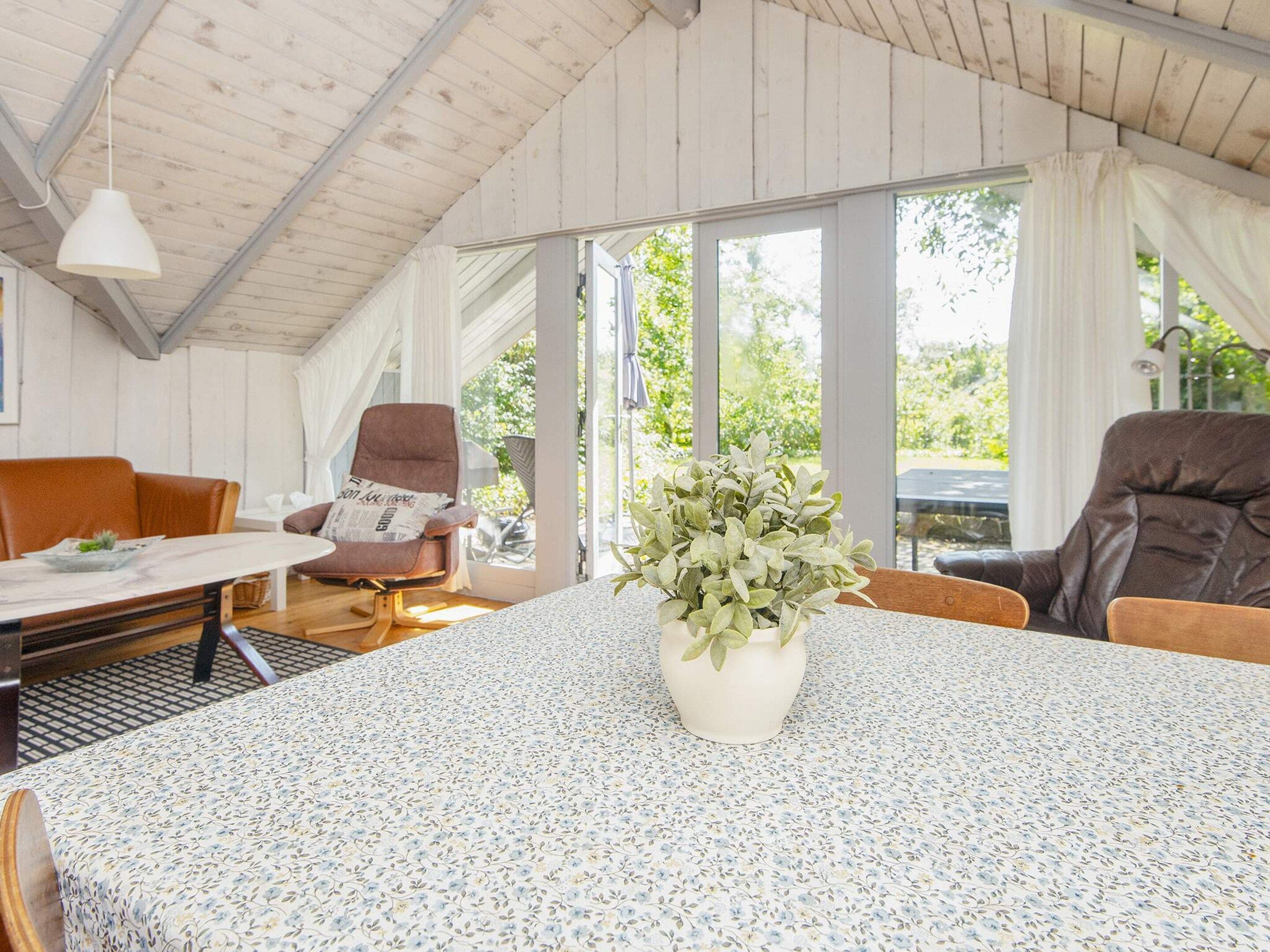 A room with a high ceiling, a round table with a floral tablecloth, two brown leather chairs, a potted plant, and a window with curtains.