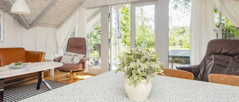 Una habitación con techo alto, una mesa redonda con mantel floreado, dos sillones de cuero marrón, una planta en maceta y una ventana con cortinas.