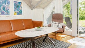 A cozy living room with a brown leather sofa, a round white table, and a patterned rug. There are two abstract paintings on the wall, a white pendant light, and a newspaper cushion on a chair. - 4 Person Holiday Home in Oksbol (Oksbol)