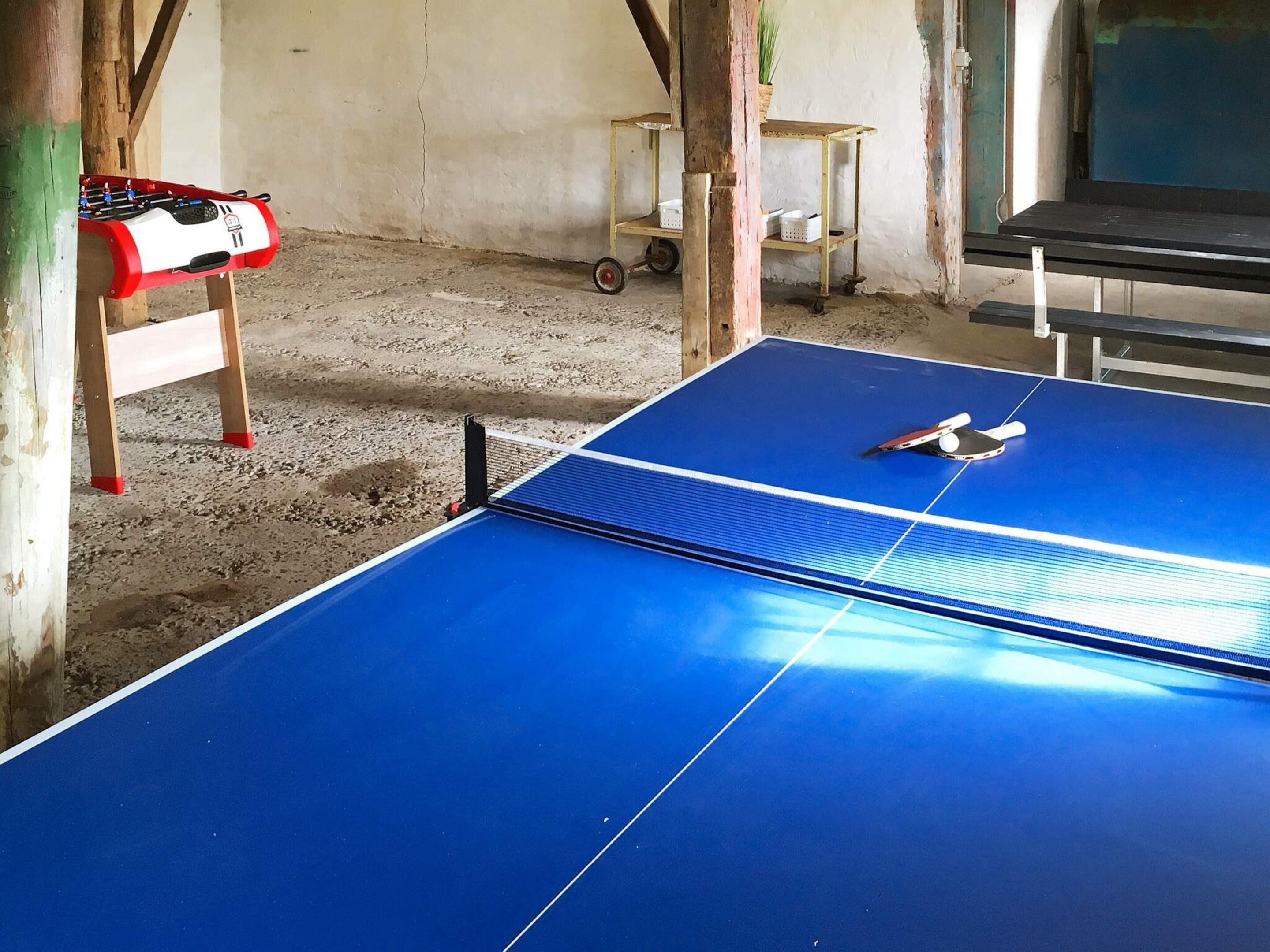 A blue ping pong table with a paddle and a foosball table in a room with a concrete floor and wooden beams.