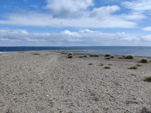 A pebbly beach with sparse vegetation and a distant view of the sea under a partly cloudy sky. - 6 Person Holiday Home in Farso (Farso)