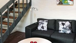 A living room with a dark grey sofa, two patterned throw pillows, a white coffee table with red candle holders, a staircase with dark railings, and a colorful abstract painting on the wall.