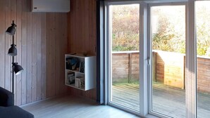 Un salon moderne avec un plancher en bois, un canapé noir, une bibliothèque blanche remplie de livres et une porte-fenêtre coulissante donnant sur un espace extérieur.