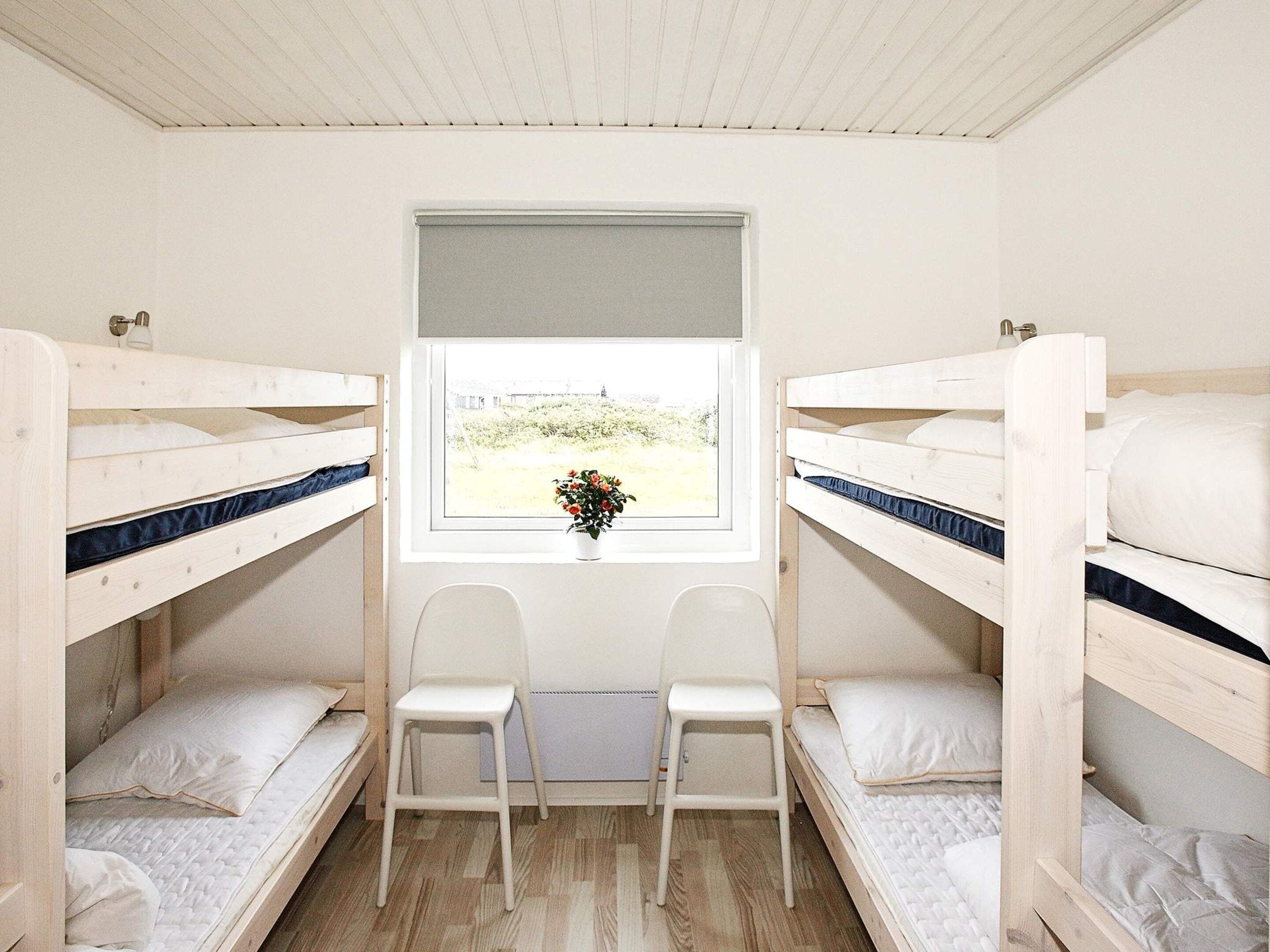 A room with bunk beds, two white chairs, a window with a view of greenery, and a small potted plant on the windowsill.