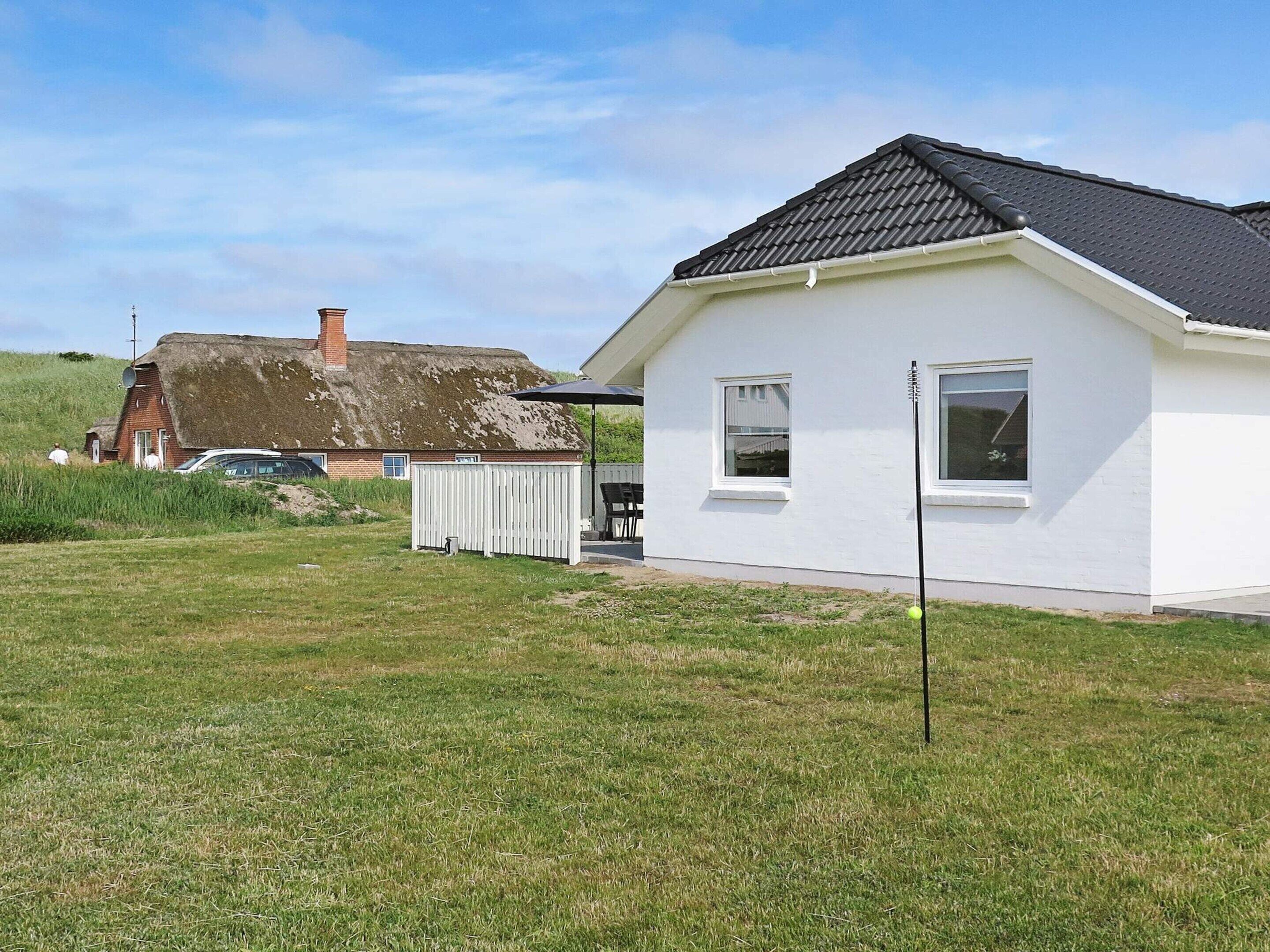 A white house with a black roof, a fenced yard, and a grassy field.