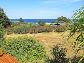 A coastal landscape with a grassy field, dense shrubbery, and a view of the sea and sailboats.