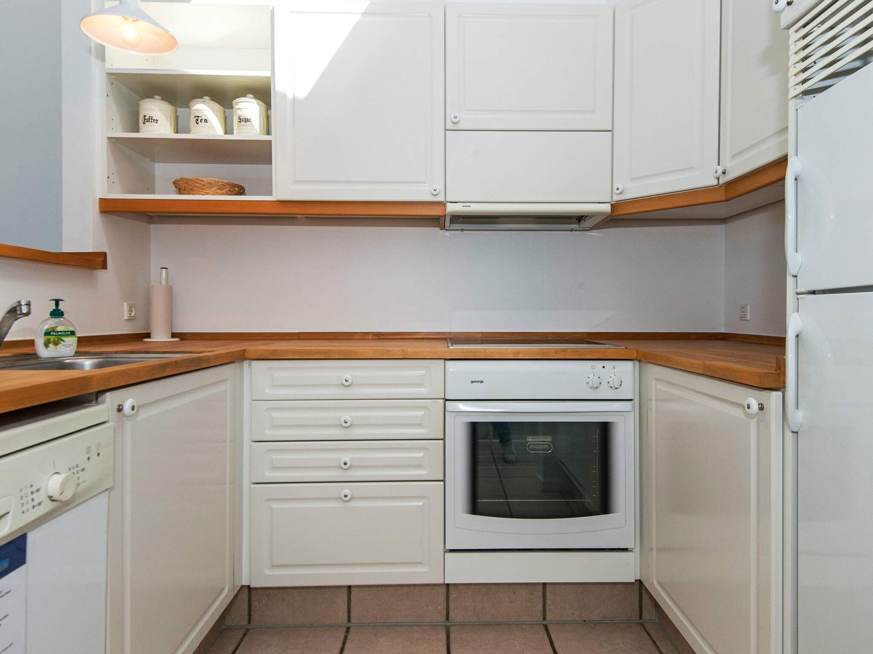 A compact kitchen with white cabinets, a wooden countertop, a built-in oven, and a shelf with jars.