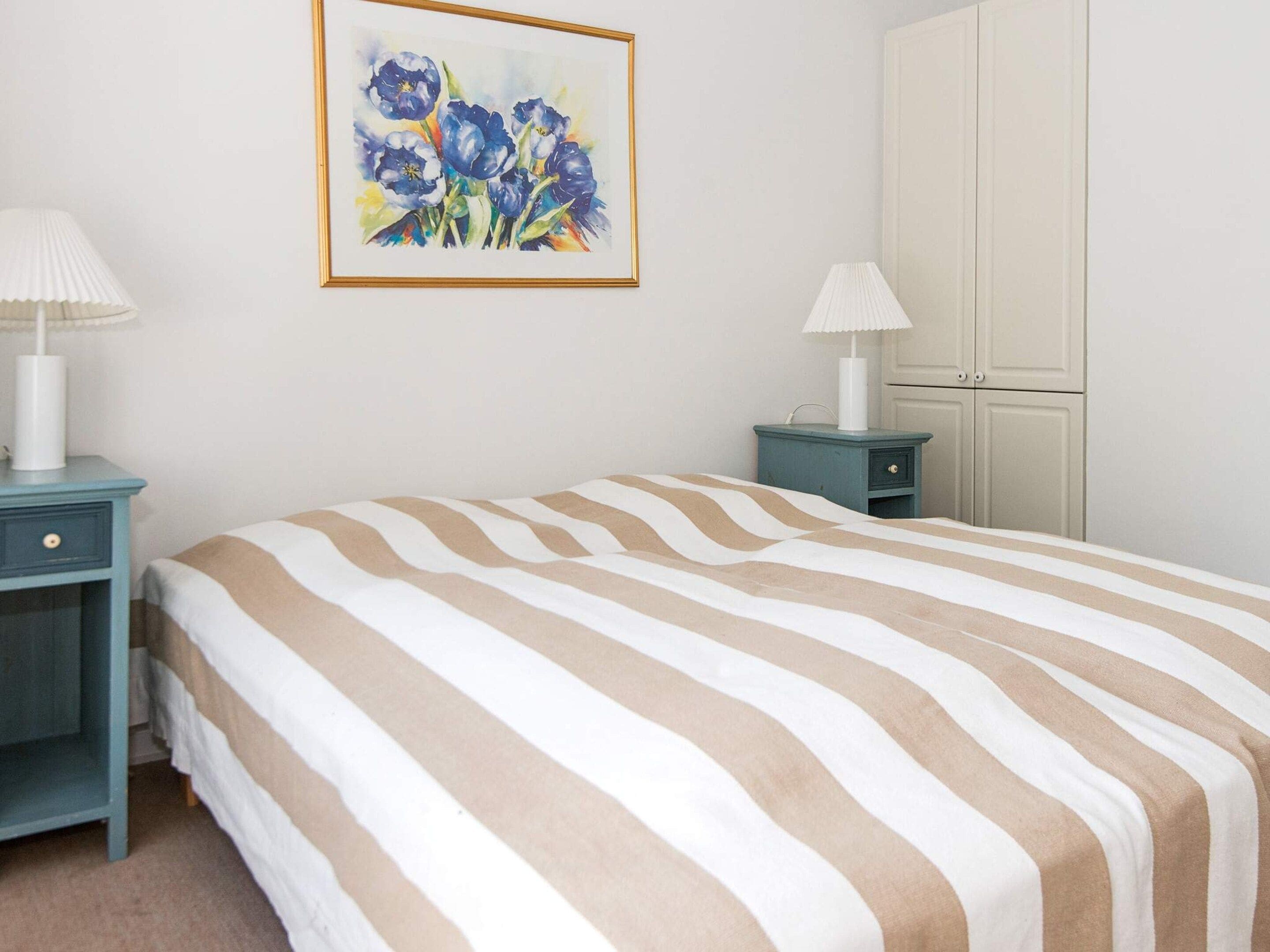 A bedroom with a striped bedspread, a nightstand with a lamp, a dresser, and a framed painting on the wall.