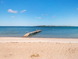A sandy beach with a wooden pier extending into calm waters, a clear sky, and a distant landmass.