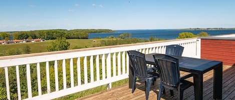 A wooden deck with a white railing, two chairs, and a table overlooking a body of water and distant landscape.