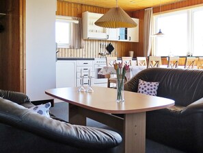 A cozy living area with a wooden table, a sofa, and a dining table with chairs. There is a kitchen in the background with white cabinets and a hanging light fixture. - 8 Person Holiday Home in Harboore (Harboore)