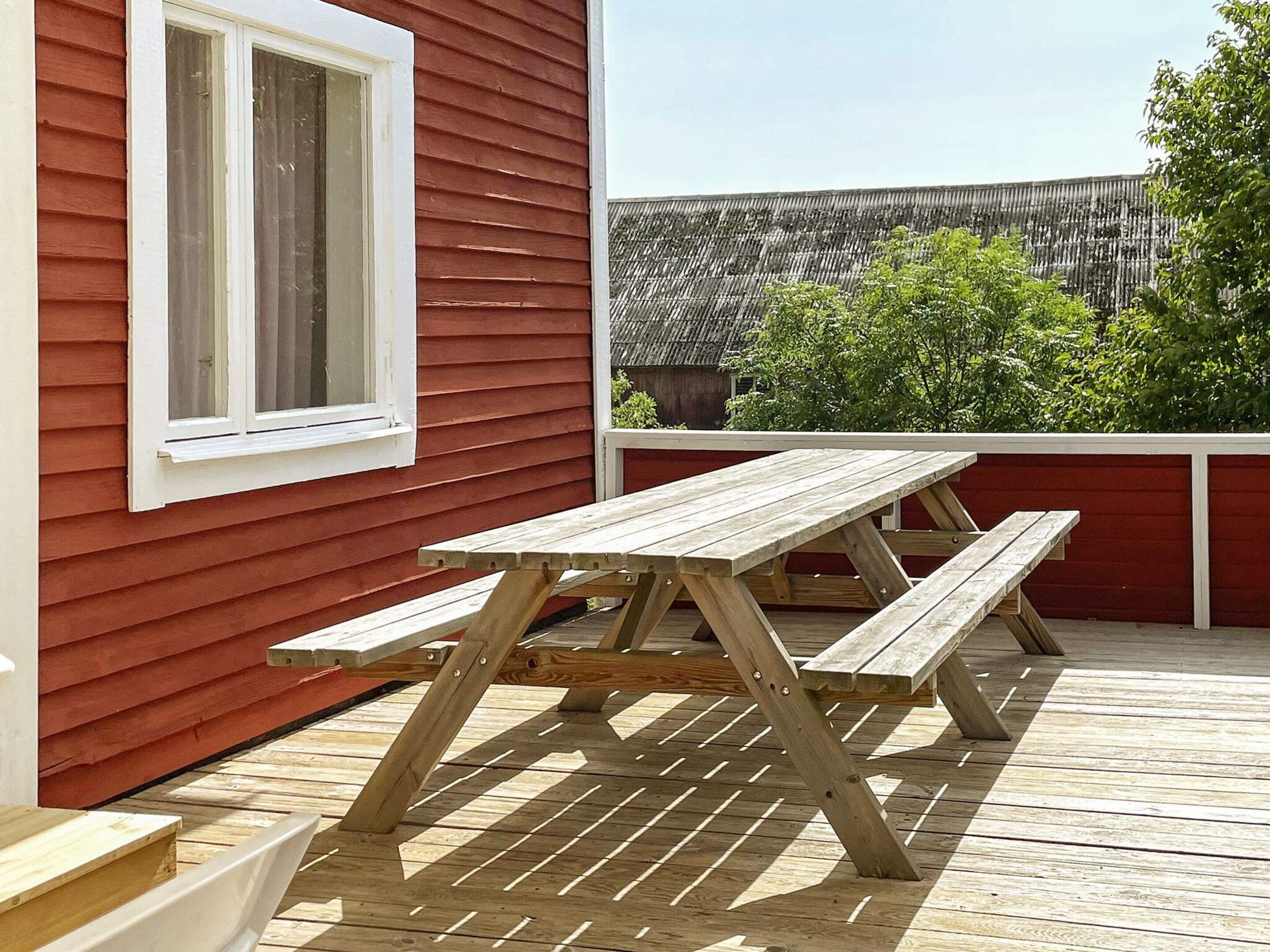 A wooden deck with a picnic table and benches, a window, and a view of a fence and greenery.