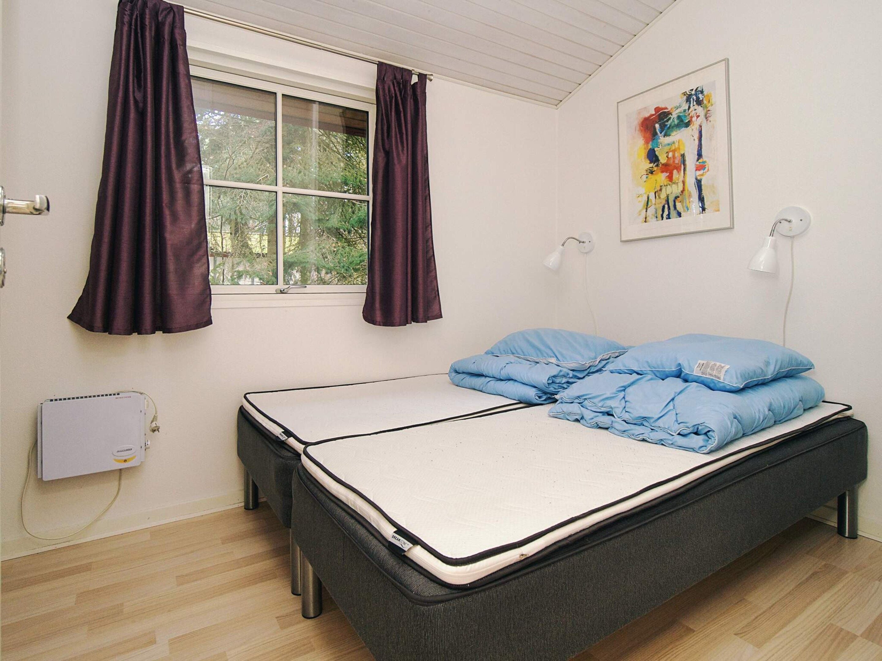 A single bed with a mattress, two pillows, a window with curtains, a wall-mounted lamp, a radiator, and a painting.