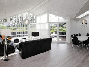 A modern living space with a black sofa, a glass coffee table, a TV, and a dining area with a white table and black chairs. - 5 Star Holiday Home in Lokken (Lokken)