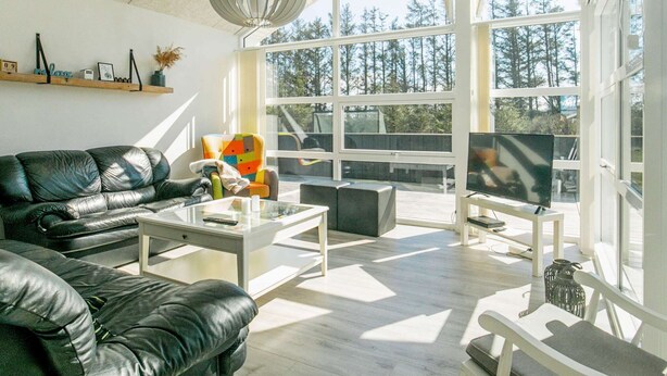 A modern living room with a black leather sofa, a colorful armchair, a glass coffee table, and a television on a white stand. Large windows offer a view of trees and a deck outside.