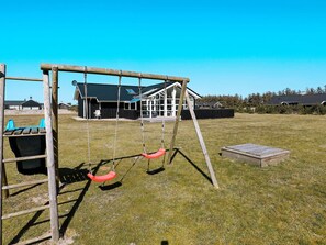 A playground with swings and a slide, a wooden bench, and a building with a dark roof in the background.