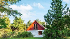 Une cabane avec un toit rouge et des murs recouverts de bardage blanc, entourée d’une forêt dense et d’une cour gazonnée.
