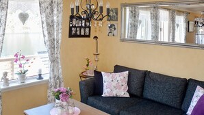 Een gezellige woonkamer met een bank, een bijzettafeltje, een kroonluchter en een groot raam met gordijnen.