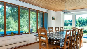 A dining area with a large table and chairs, large windows with a view of greenery, and a ceiling fan.