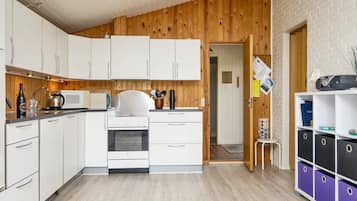 A compact kitchen with white cabinets, a stove, and a microwave. There's a wooden wall paneling, a door leading to another room, and a shelf with storage boxes.