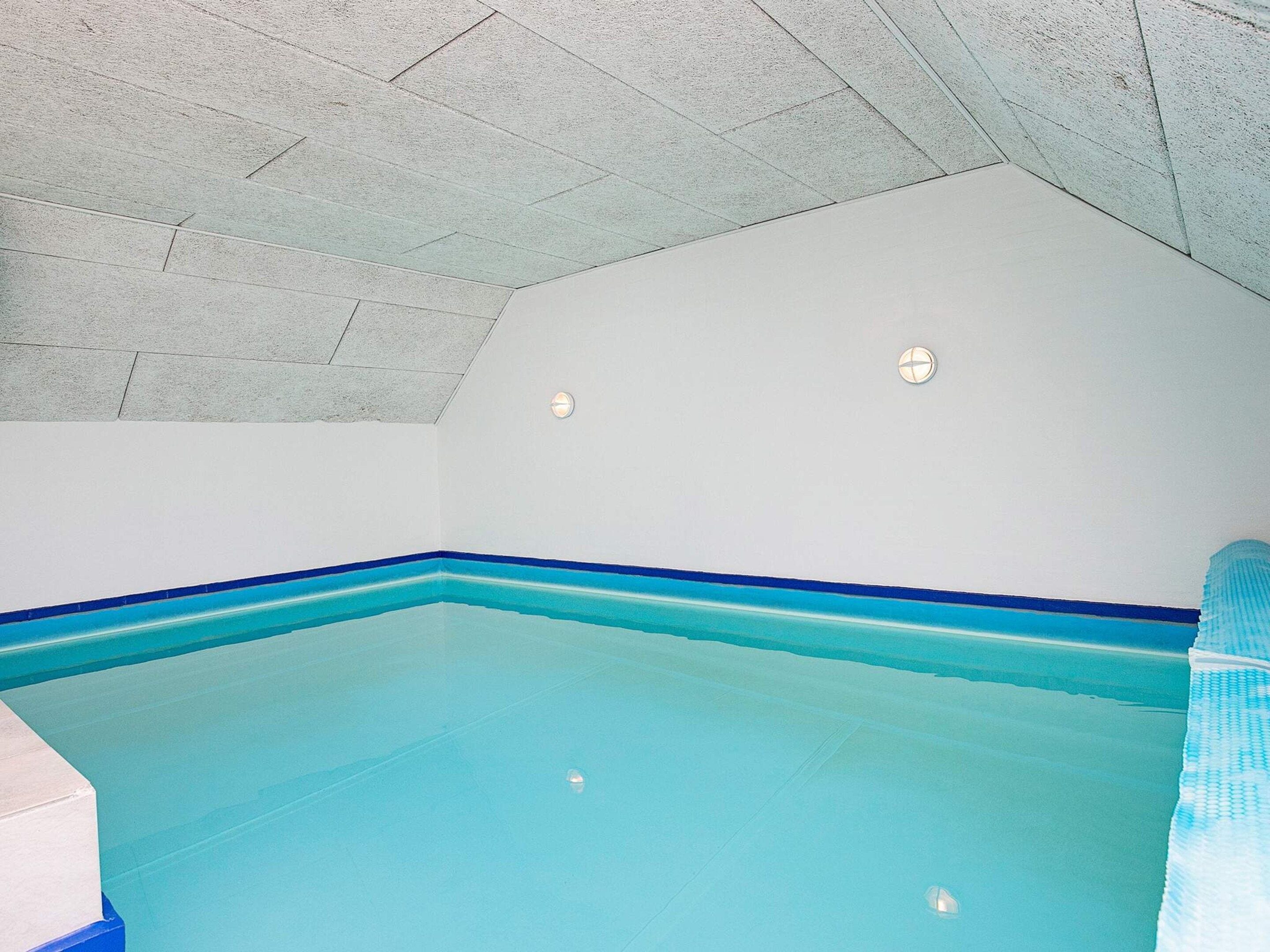 A swimming pool with a blue edge, a ceiling with a herringbone pattern, and a white wall with two light fixtures.
