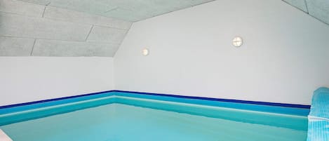 A swimming pool with a blue edge, a ceiling with a herringbone pattern, and a white wall with two light fixtures.