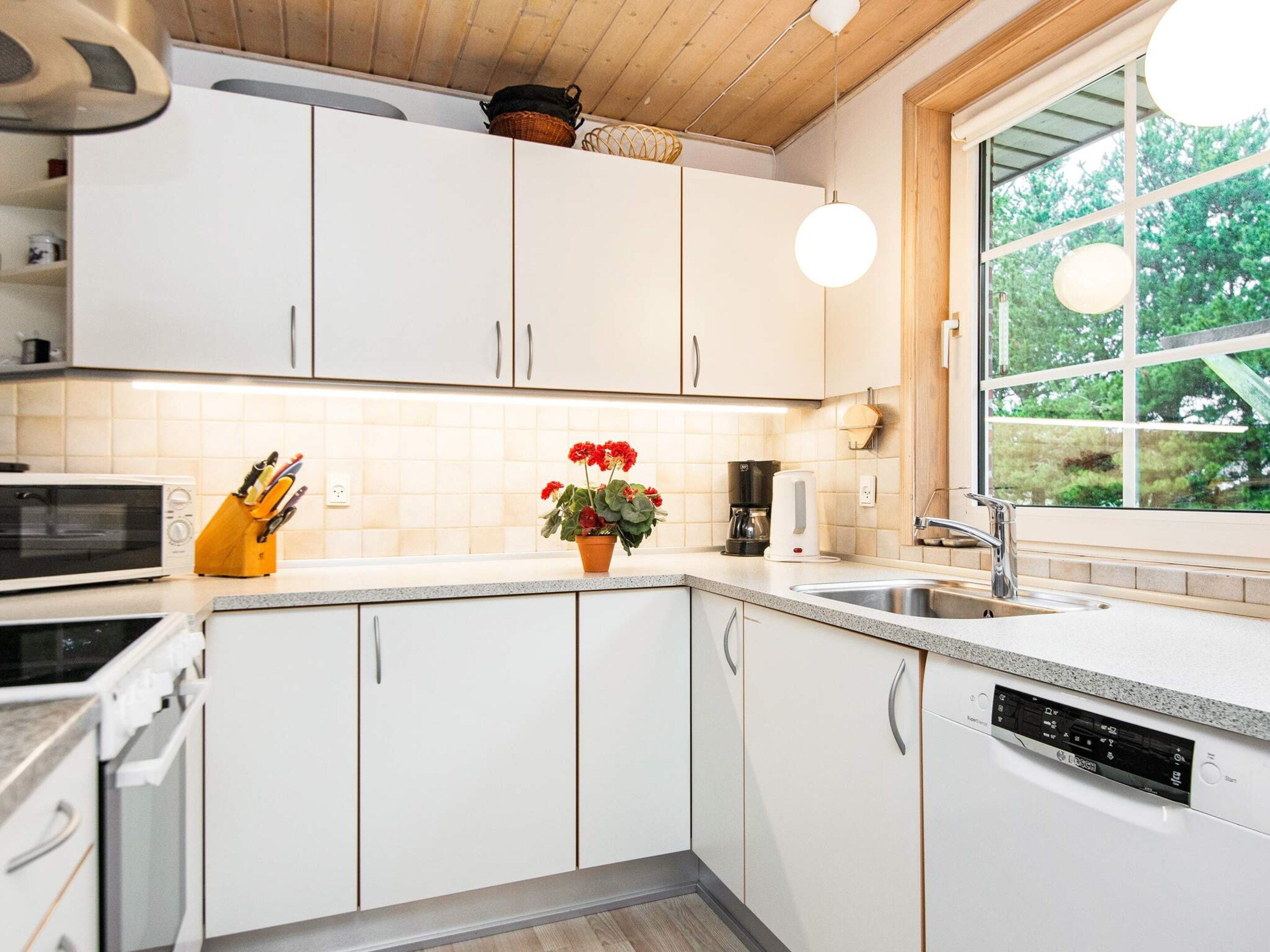 A modern kitchen with white cabinetry, a built-in microwave, a coffee maker, and a window with a view of trees.