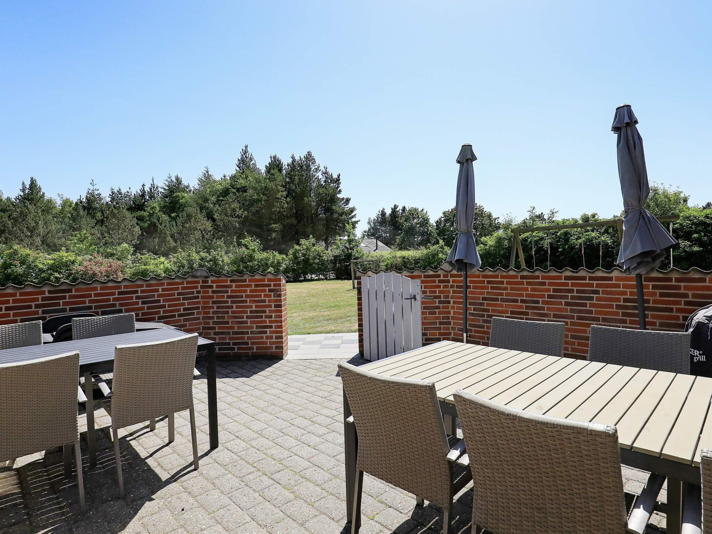 En udendørs patio med murstensvægge, et træbord, fletstole og en grill.