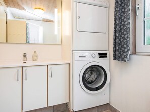 A compact laundry room with a front-loading washing machine, a small cabinet with a mirror, and a window with a patterned curtain. - 4 Star Holiday Home in Oksbol (Oksbol)
