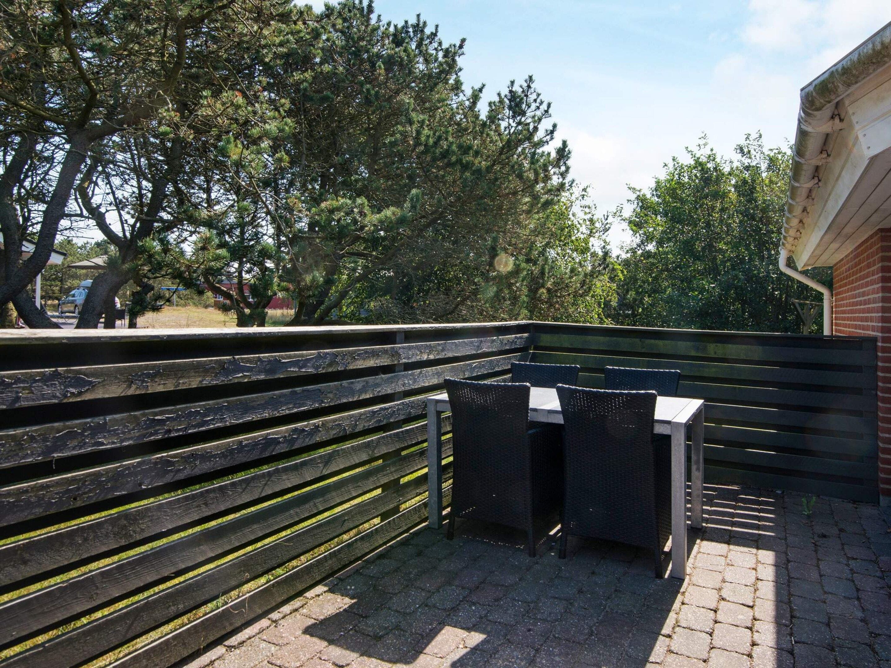 An outdoor seating area with a wooden bench and chairs, surrounded by trees and a brick wall.