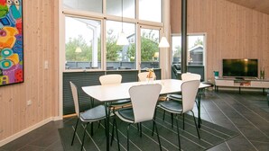 Un comedor con una mesa blanca y sillas negras, ventanas amplias y un televisor sobre un mueble.