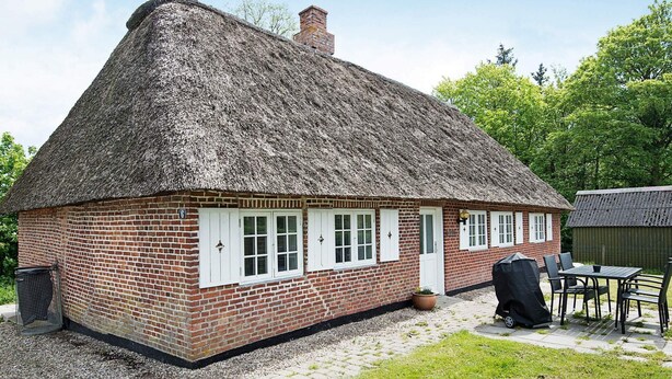 Une maison en briques avec un toit de chaume, des volets blancs et un petit espace extérieur aménagé avec une table et des chaises.