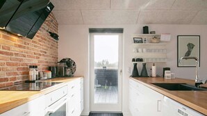 Cozinha moderna com armários brancos, bancada de madeira e parede de tijolos. Há uma porta que dá para uma varanda, e vários utensílios de cozinha à vista no balcão.