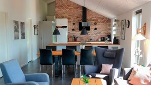 Uma sala de estar moderna com uma mesa de centro de madeira, sofás azuis e uma cozinha ao fundo, com parede de tijolos e luminárias pendentes.