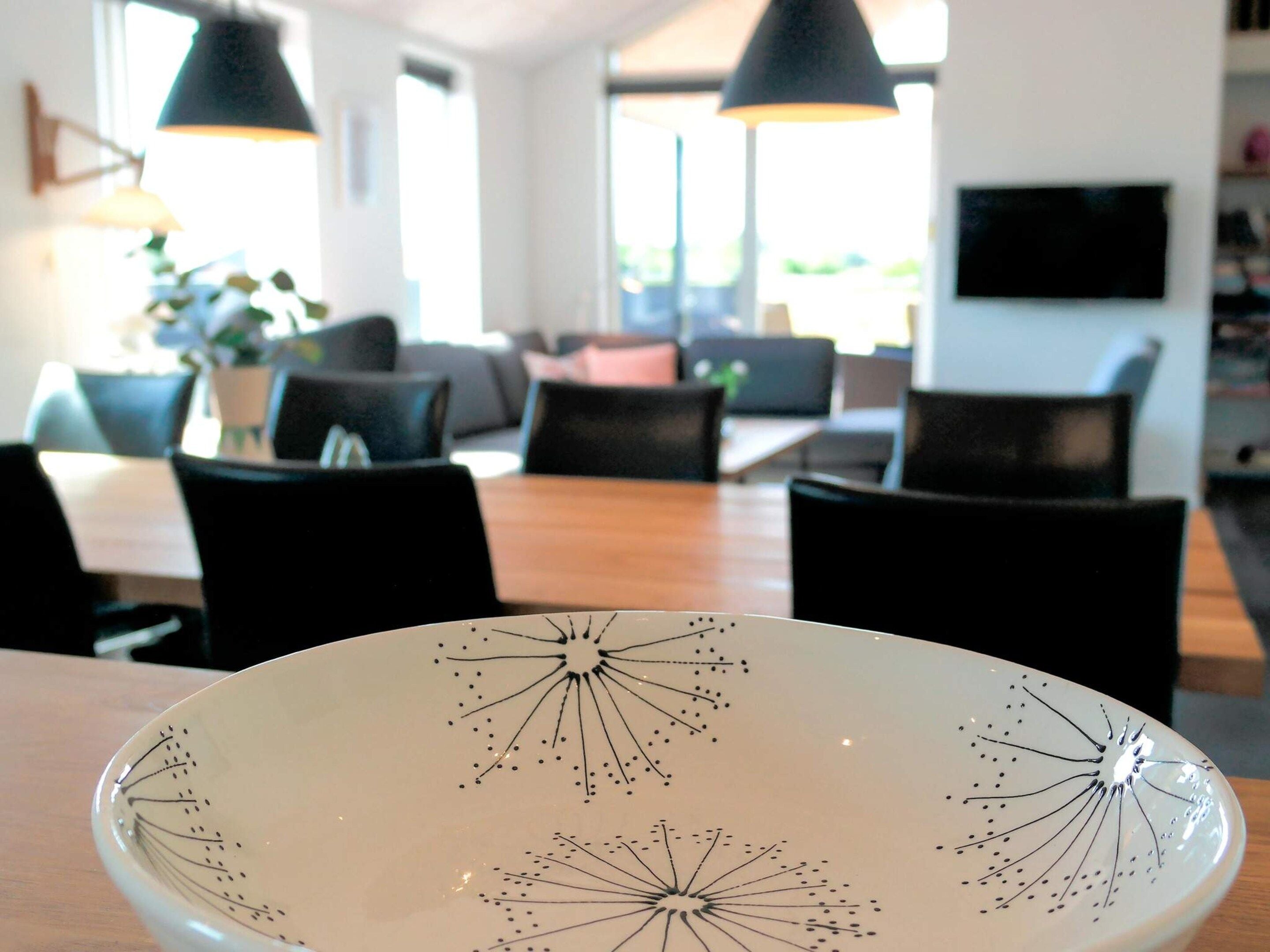 Un comedor moderno con un plato blanco con diseño geométrico, sillas negras, una mesa de madera y un ventanal grande que proporciona luz natural.
