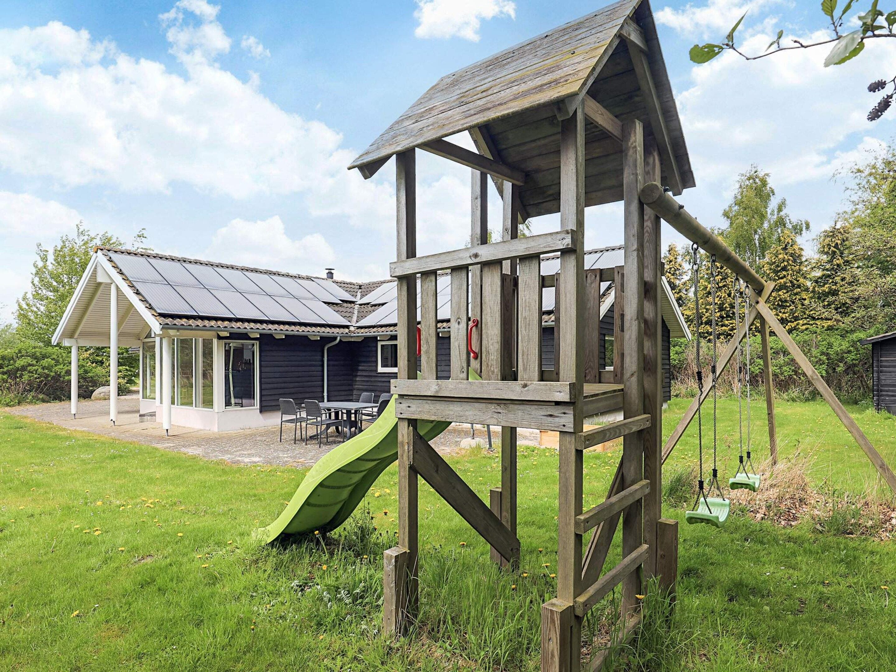 A wooden playhouse with a slide, a covered seating area, and a house with a sloped roof in the background.