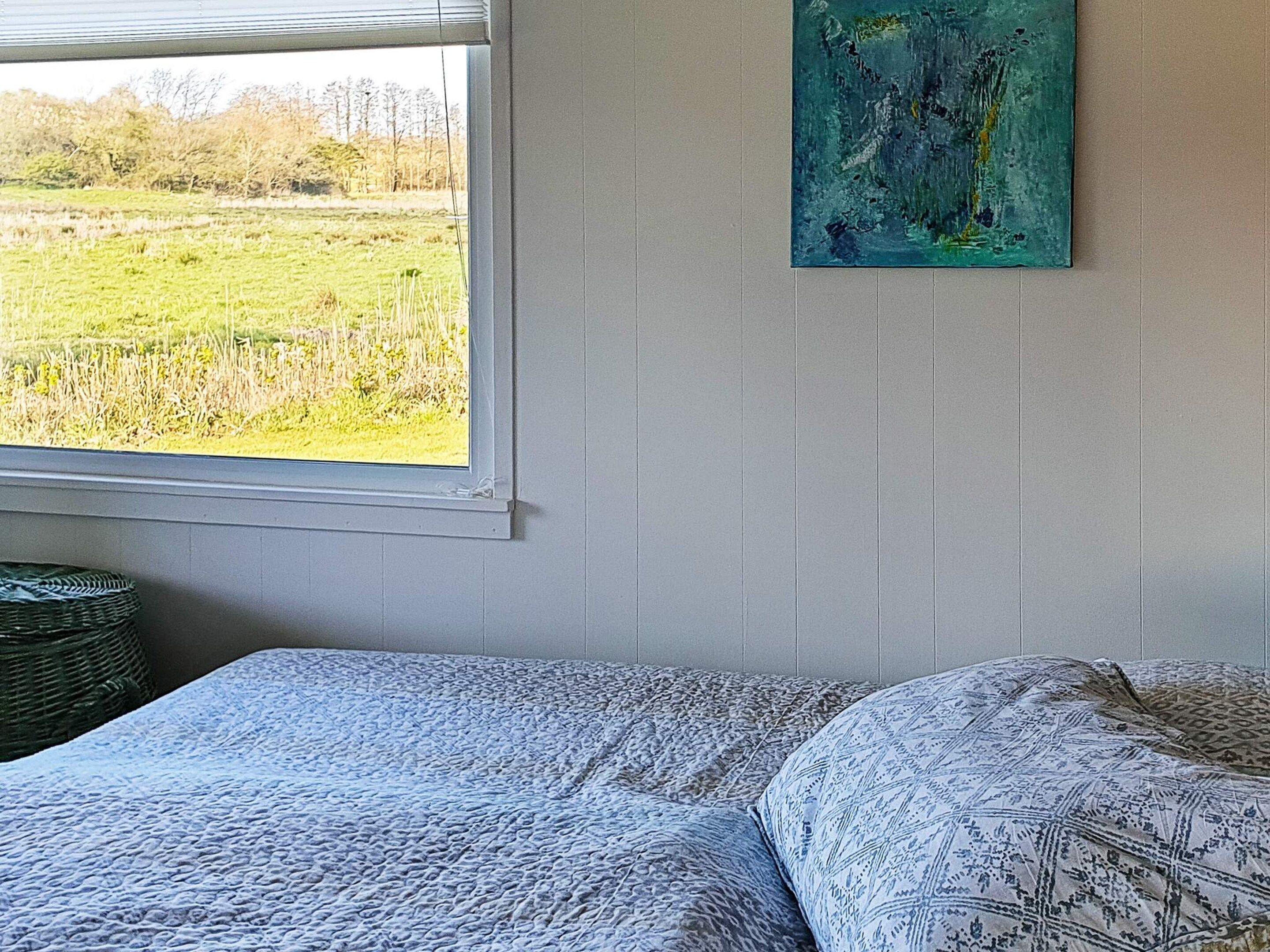 A room with a bed, a window with a view of a field, and a painting on the wall.