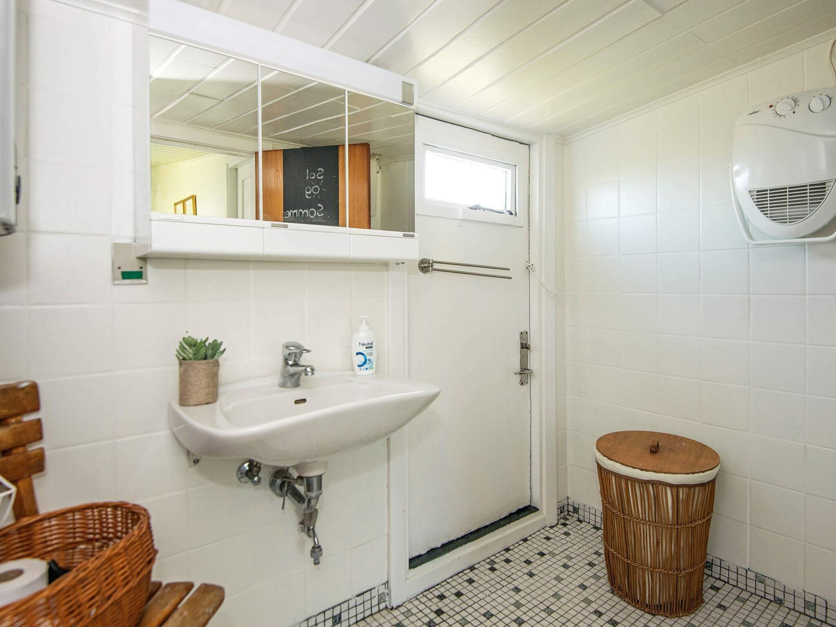Une salle de bain avec un lavabo blanc, un miroir, une petite plante en pot, une bouteille blanche, un panier en osier et un tabouret rond en osier.
