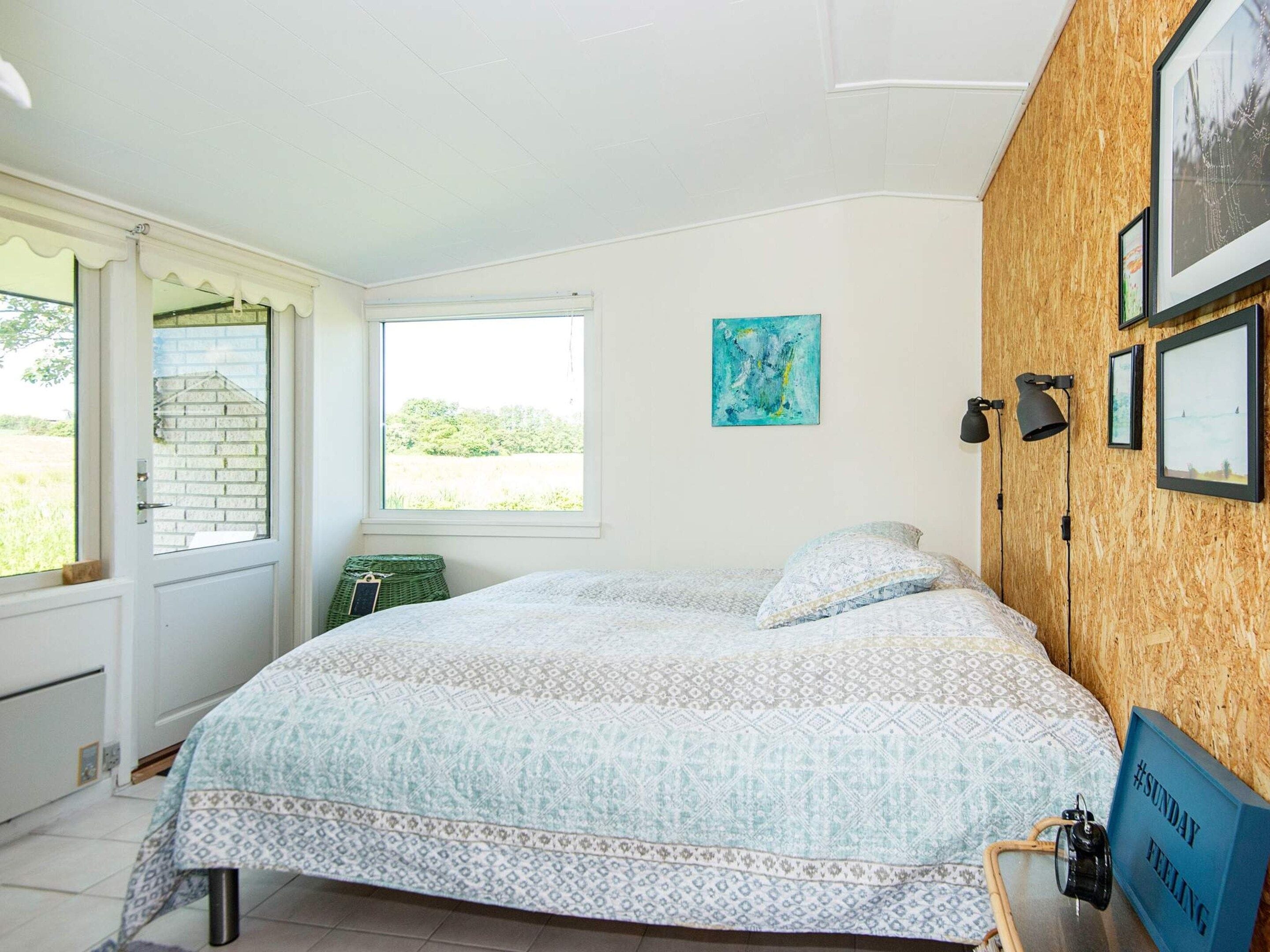 A bedroom with a bed, a window, a wall-mounted picture, and a small table with a lamp.