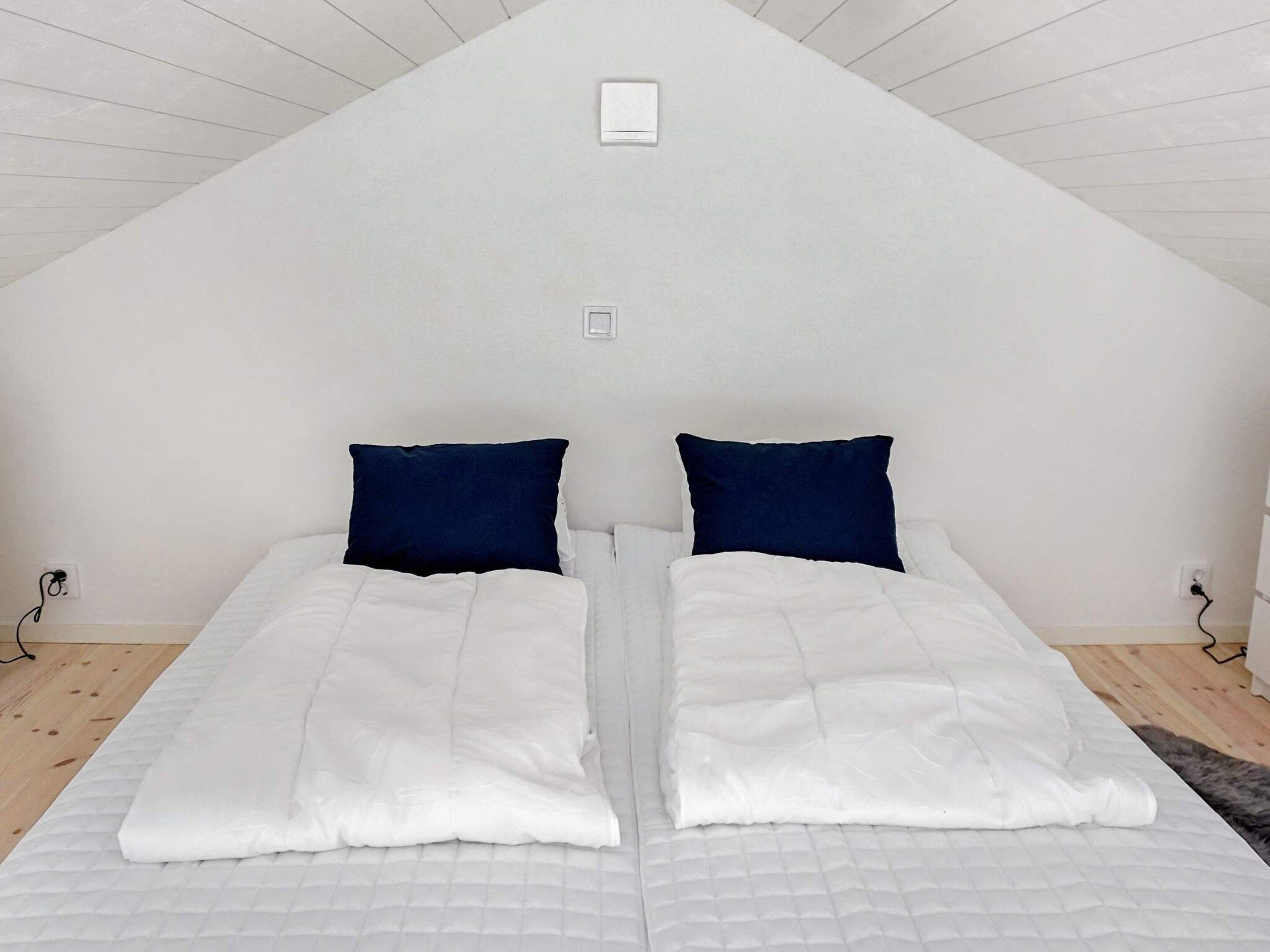 A bedroom with a sloped ceiling, two beds with white bedding and dark blue pillows, and a small white light fixture above the beds.