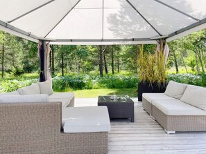 A covered outdoor seating area with wicker furniture and a glass table, surrounded by lush greenery. - Holiday Home in Katthammarsvik (Katthammarsvik)