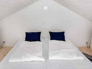 Une pièce avec un plafond en pente, deux lits avec une literie blanche et des oreillers bleu foncé, et un petit luminaire blanc au-dessus des lits.