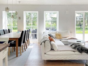 A modern living room with a white sofa, a dining table with chairs, and large windows offering a view of greenery. - Holiday Home in Katthammarsvik (Katthammarsvik)