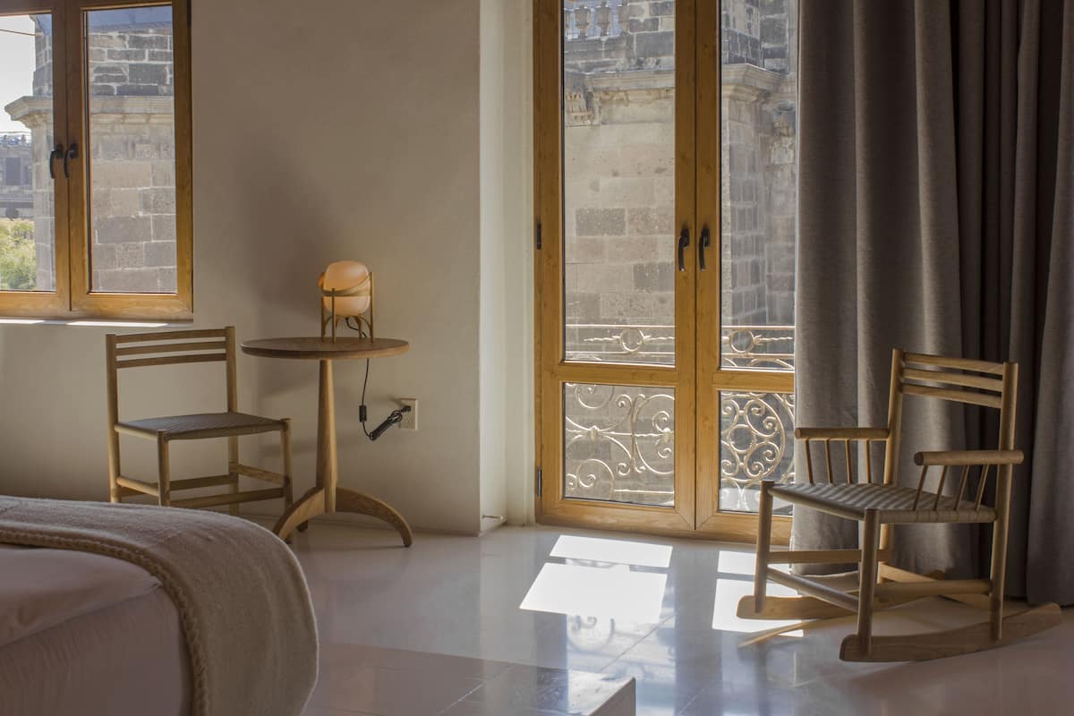 A room with a bed, two wooden chairs, a small table, and a window with a view of a stone building.
