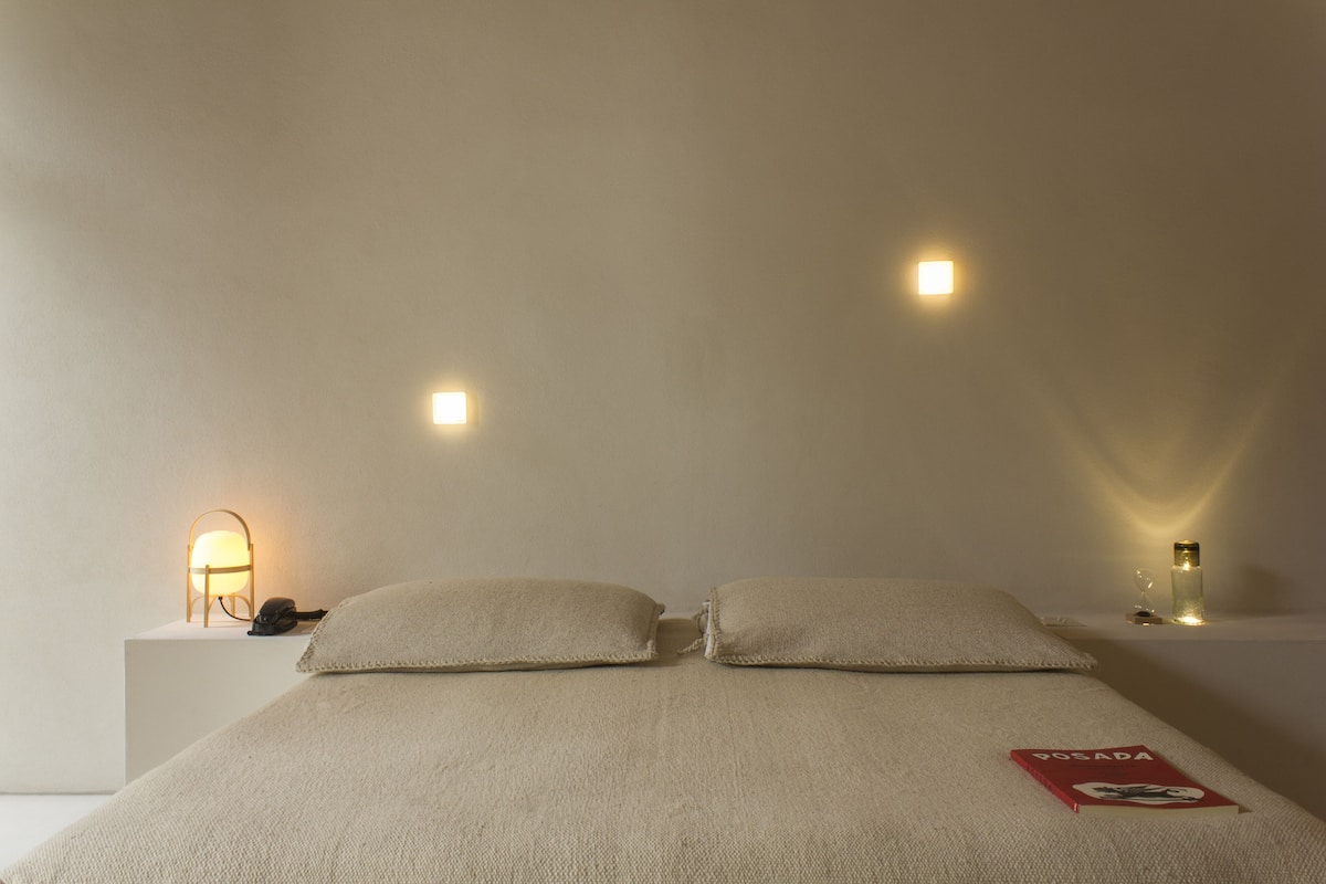 A neatly made bed with pillows, a bedside table with a lamp, and a book titled "POSADA" on the bed.