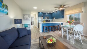 Un salon moderne avec un canapé bleu foncé, une table basse en bois sur laquelle est posé un panier de fruits, et un coin repas avec des chaises en osier blanc et une table en verre.