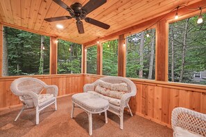 A wooden deck with wicker furniture, a ceiling fan, and large windows overlooking a forest. - The Swiss Chalet | Mount Gretna (Mount Gretna)