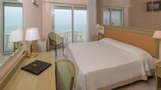 Ein Hotelzimmer mit einem großen Bett, einem Schreibtisch mit Stuhl, einem kleinen Tisch und einem Fenster mit Meerblick.