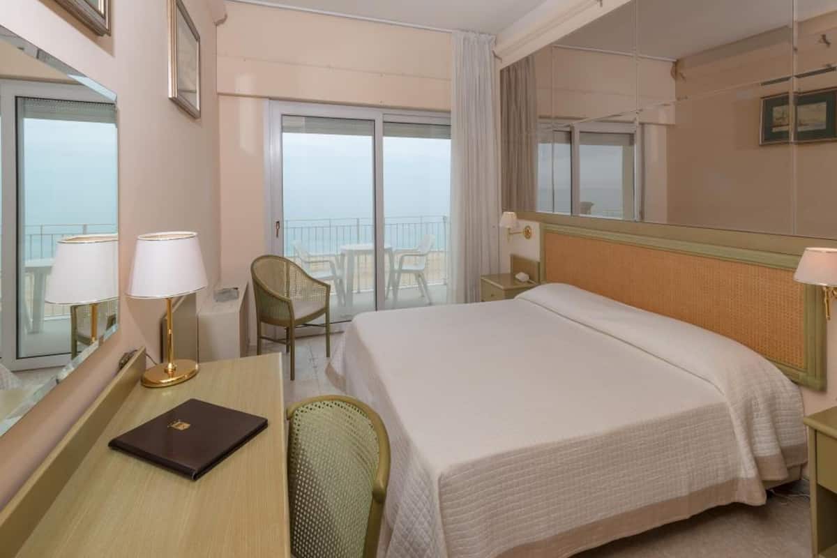 A hotel room with a large bed, a desk with a chair, a small table, and a window with a view of the sea.